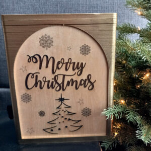 A wine box next to a christmas tree. The box has a gold frame and an engraved oak side panel that says "Merry Christmas".