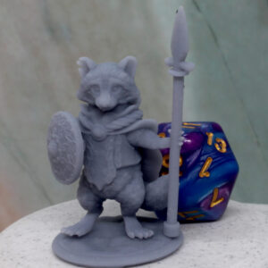 A tabletop miniature of a raccoon folk character holding a spear and shield, standing next to a 20 sided dice.